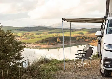 Wohnmobil mit Vorzelt und zwei Stühlen an einem See, mit Blick auf grüne Hügel und bewölkten Himmel.