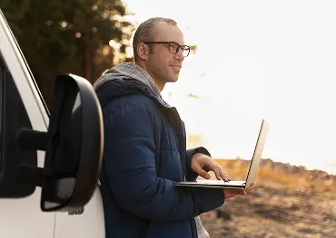 Mann mit Brille und blauer Jacke, der im Freien einen Laptop benutzt, lächelt und in die Ferne schaut.