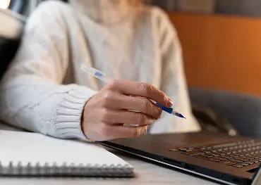 Person im weißen Pullover, die einen Stift neben einem Laptop hält, mit einem Notizbuch auf dem Tisch.
