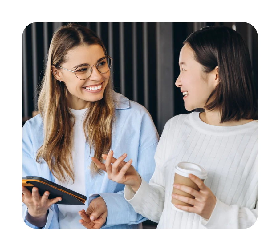 Two happy women, one with a coffee cup and the other holding a small card reader, are smiling and talking. 