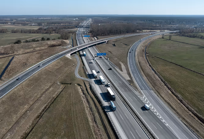 Luftaufnahme einer Autobahn mit mehreren Fahrspuren, darunter ein Überführungsviadukt und LKWs auf dem rechten Fahrstreifen, umgeben von Feldern.