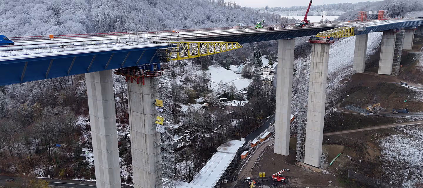 Eine hohe Brücke im Bau mit Betonpfeilern über einem schneebedeckten Tal mit Bäumen und Häusern.