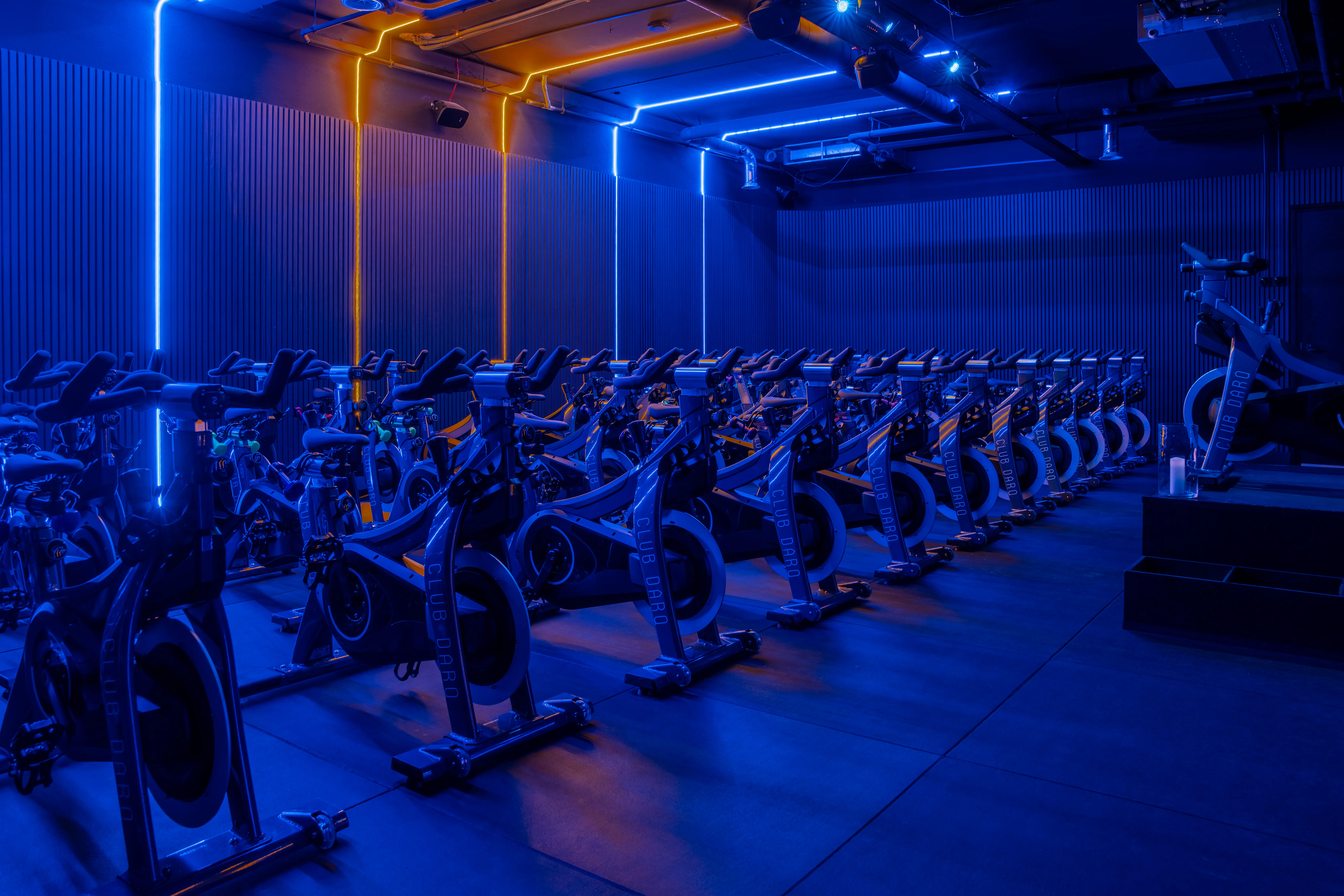 Indoor cycling studio with rows of stationary bikes illuminated by blue and orange neon lights.