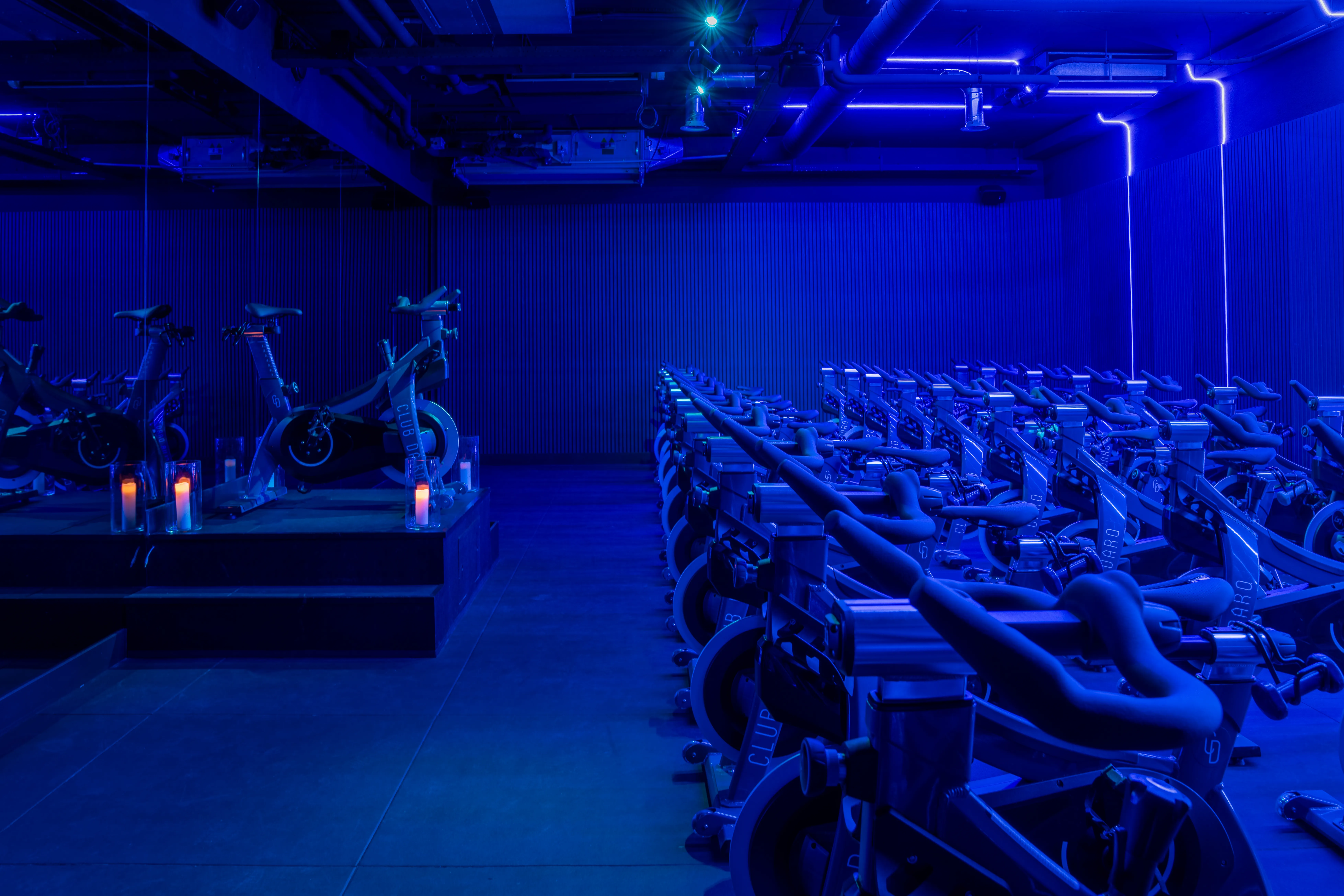 Indoor cycling studio with rows of stationary bikes under blue neon lighting and candles on a raised platform.