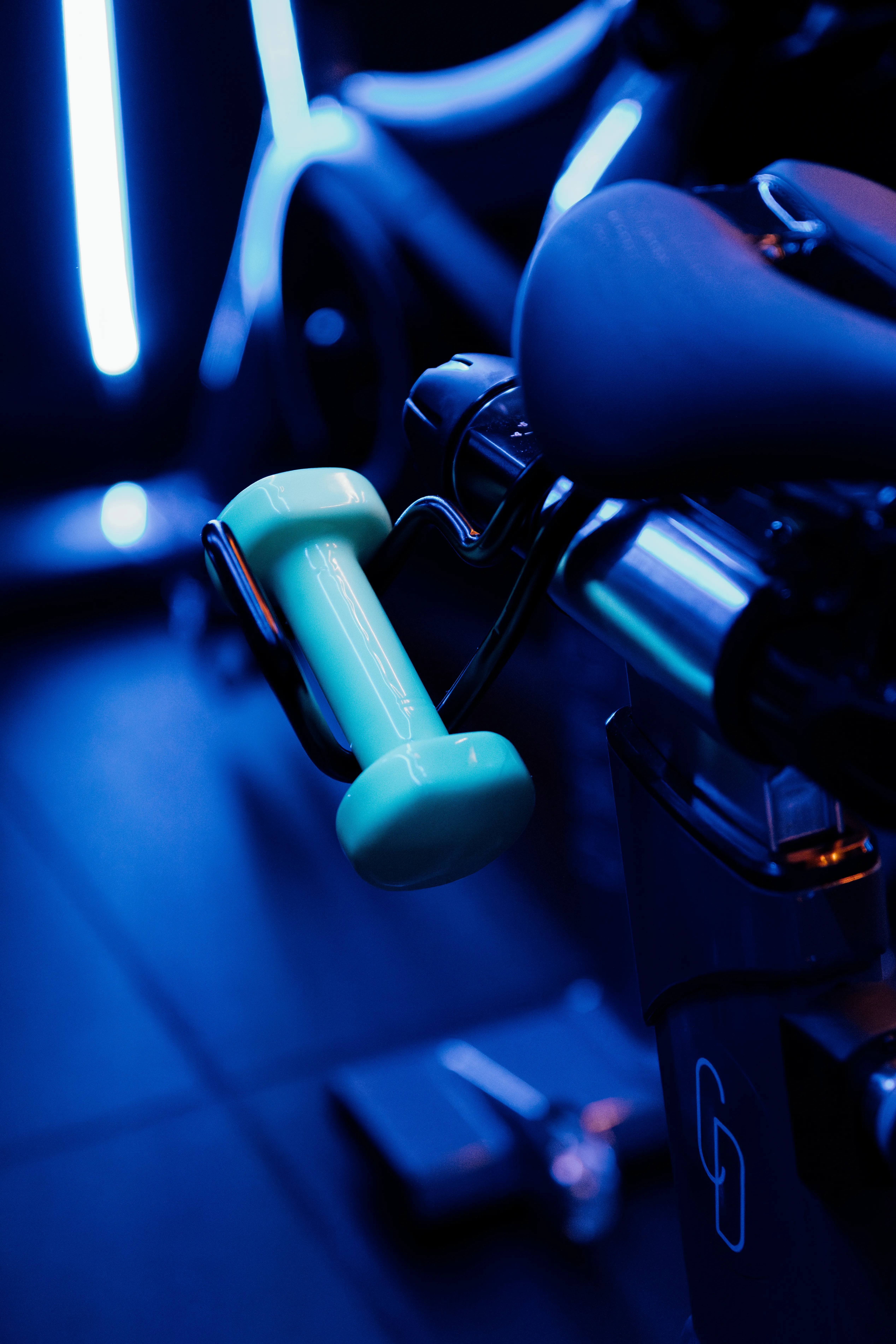 Close-up of a light blue dumbbell attached to an exercise bike handle in a gym with blue lighting.