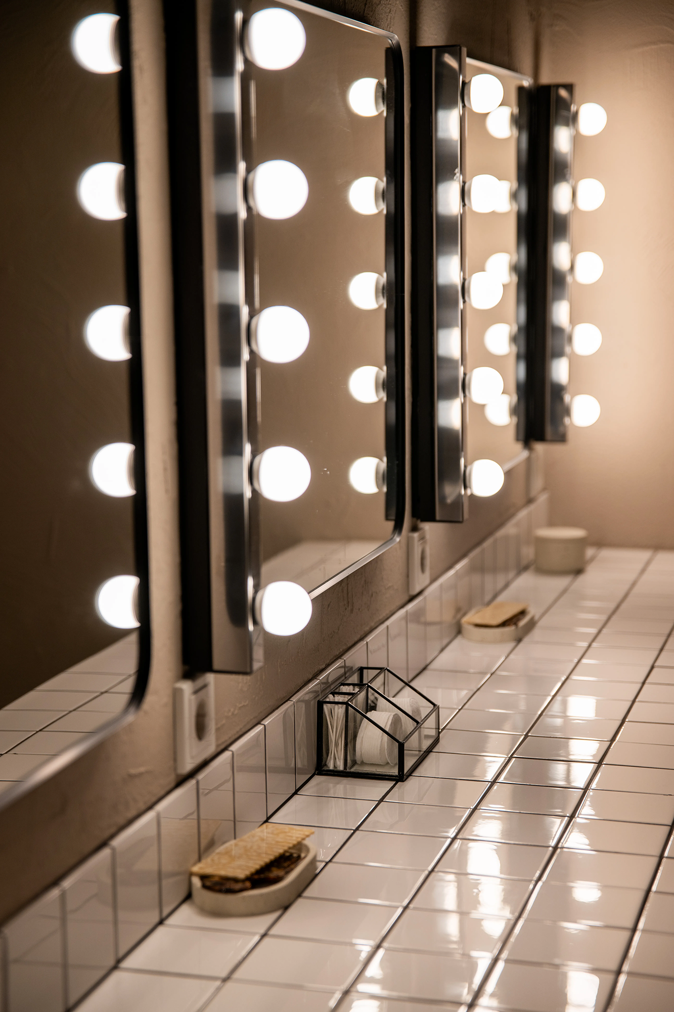 Row of illuminated vanity mirrors above a tiled counter with soap dishes and a container holding cotton pads and swabs.
