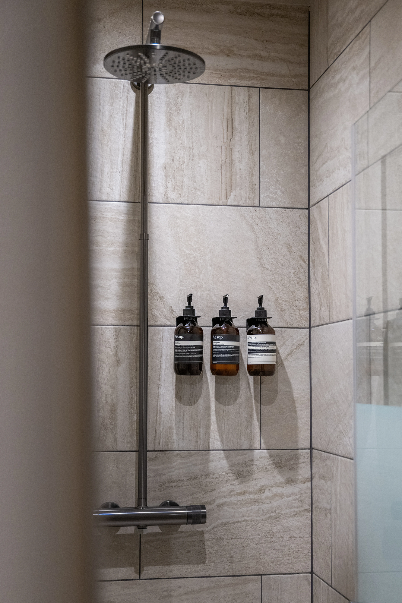 Modern shower with stainless steel rain showerhead and three amber Aesop soap dispensers mounted on beige tiled wall.
