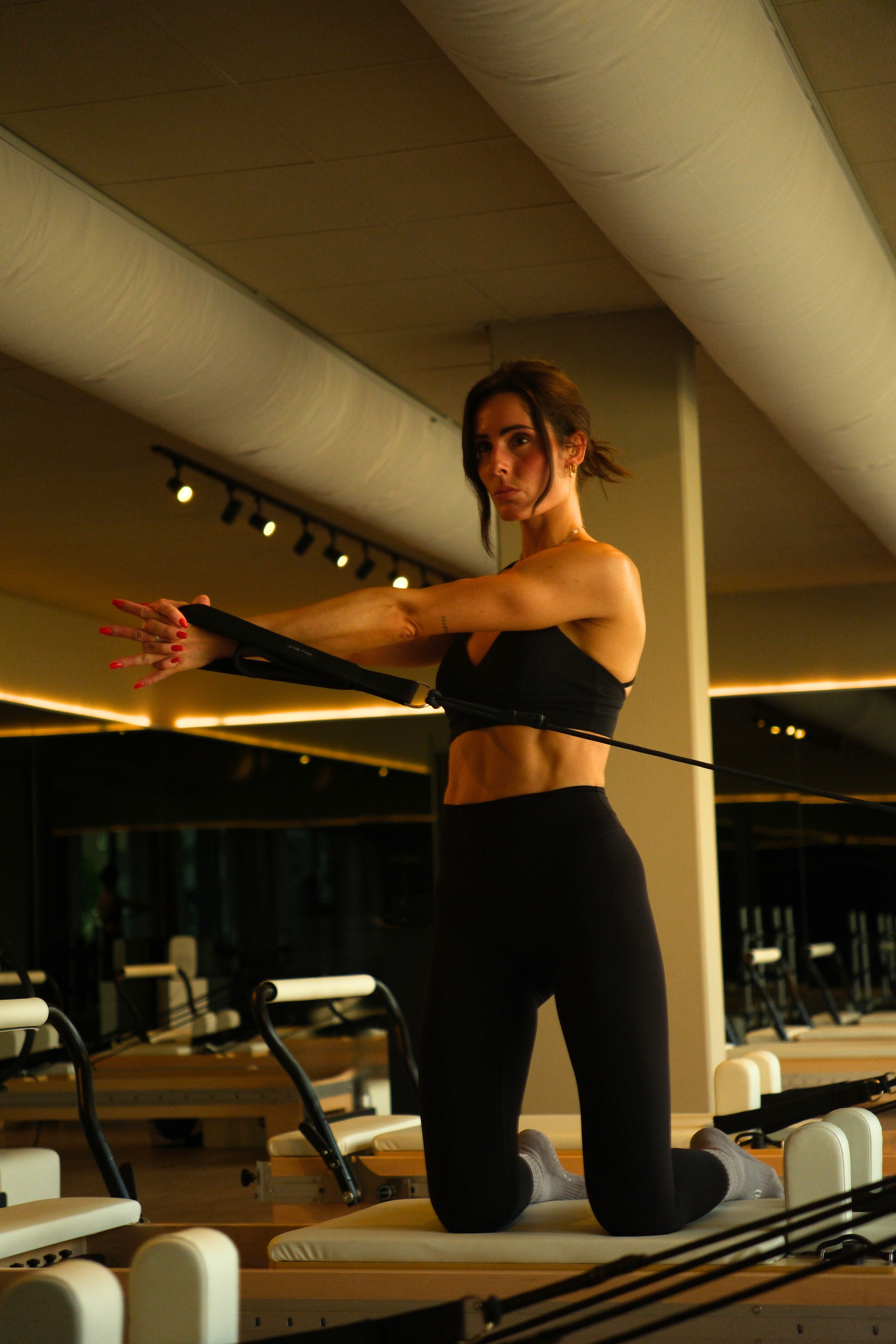 Woman in black sportswear kneeling on Pilates reformer machine while stretching arms using resistance bands in gym.