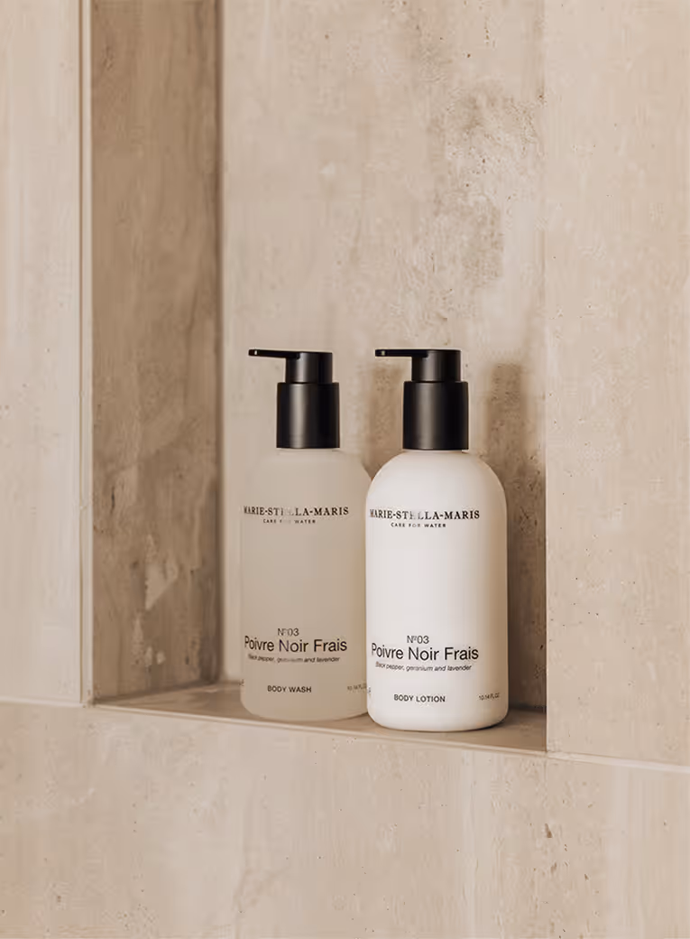 Two Marie-Stella-Maris skincare bottles with black pump dispensers, labeled Poivre Noir Frais body wash and body lotion, placed on a stone shelf.