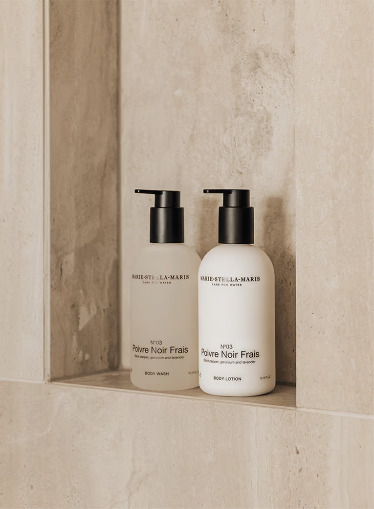 Two Marie-Stella-Maris skincare bottles with black pump dispensers, labeled Poivre Noir Frais body wash and body lotion, placed on a stone shelf.