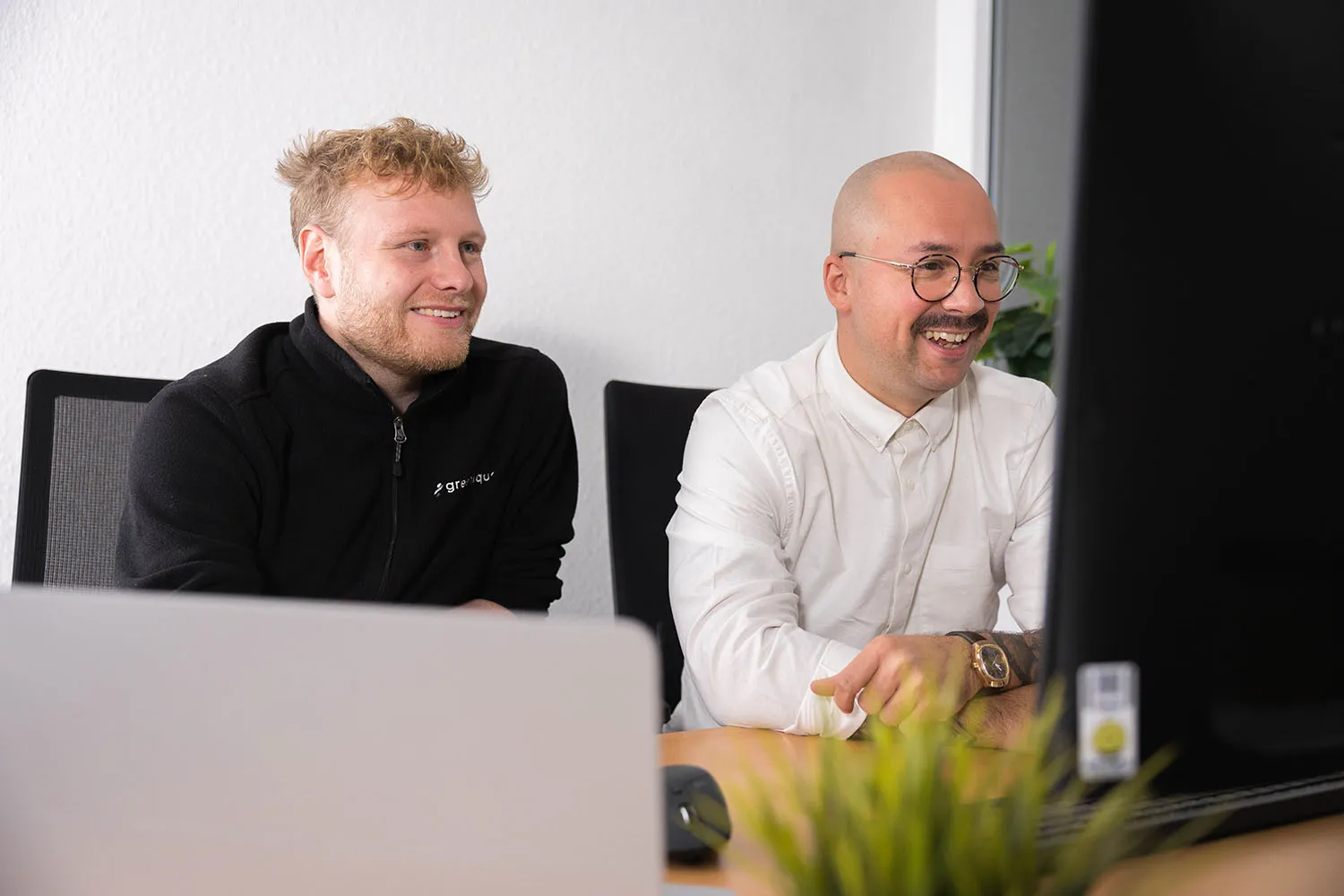 Zwei Personen sitzen lächelnd vor einem Bildschirm