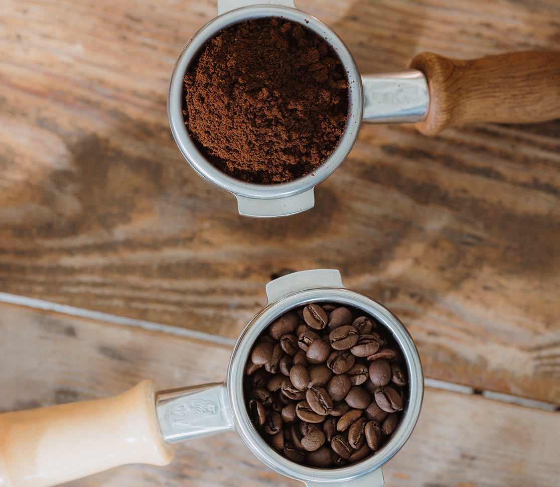 Zwei Siebträgereinsätze, einer mit Kaffeebohnen und einer mit gemahlenem Kaffee, auf einer Holzoberfläche.