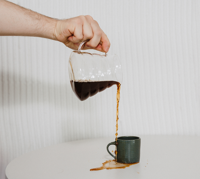 Hand pouring coffee from a glass carafe into a small green cup on a white surface, with coffee spilling over the edge.
