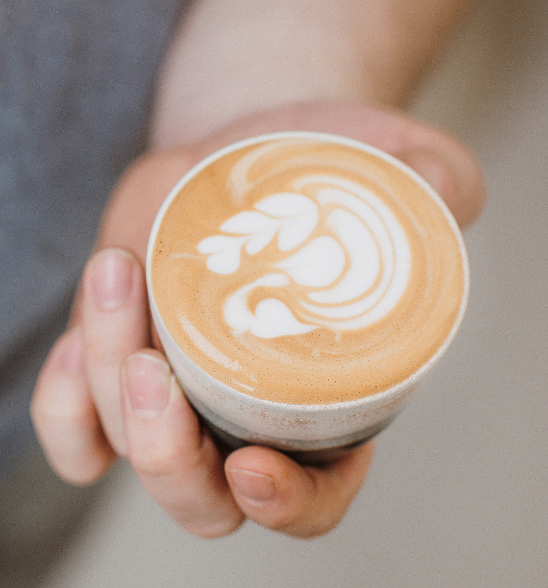 Close-up of hand holding a cup of latte with white leaf-shaped foam art on top.