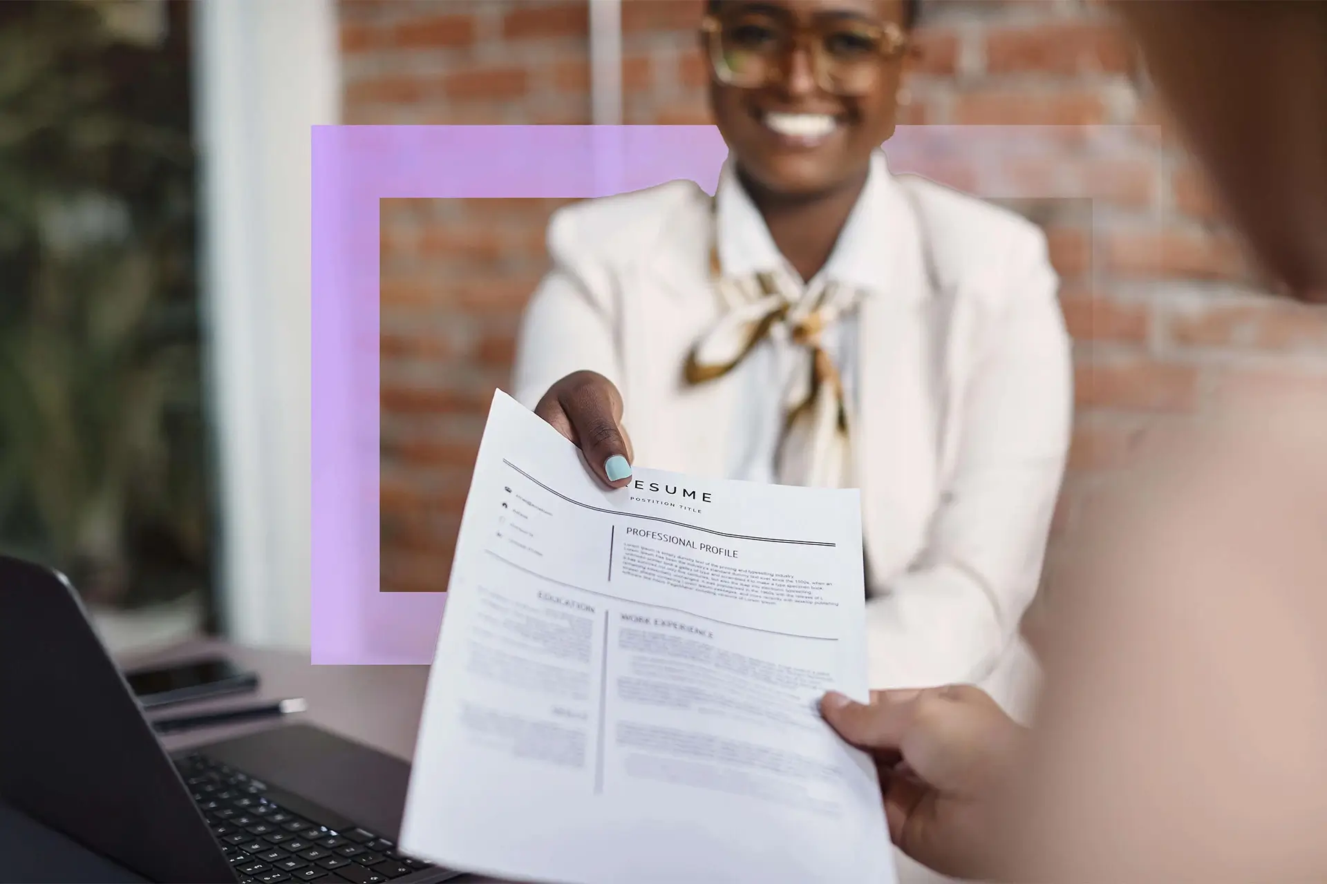 Smiling woman in a white blazer handing a resume across a desk with a laptop visible.
