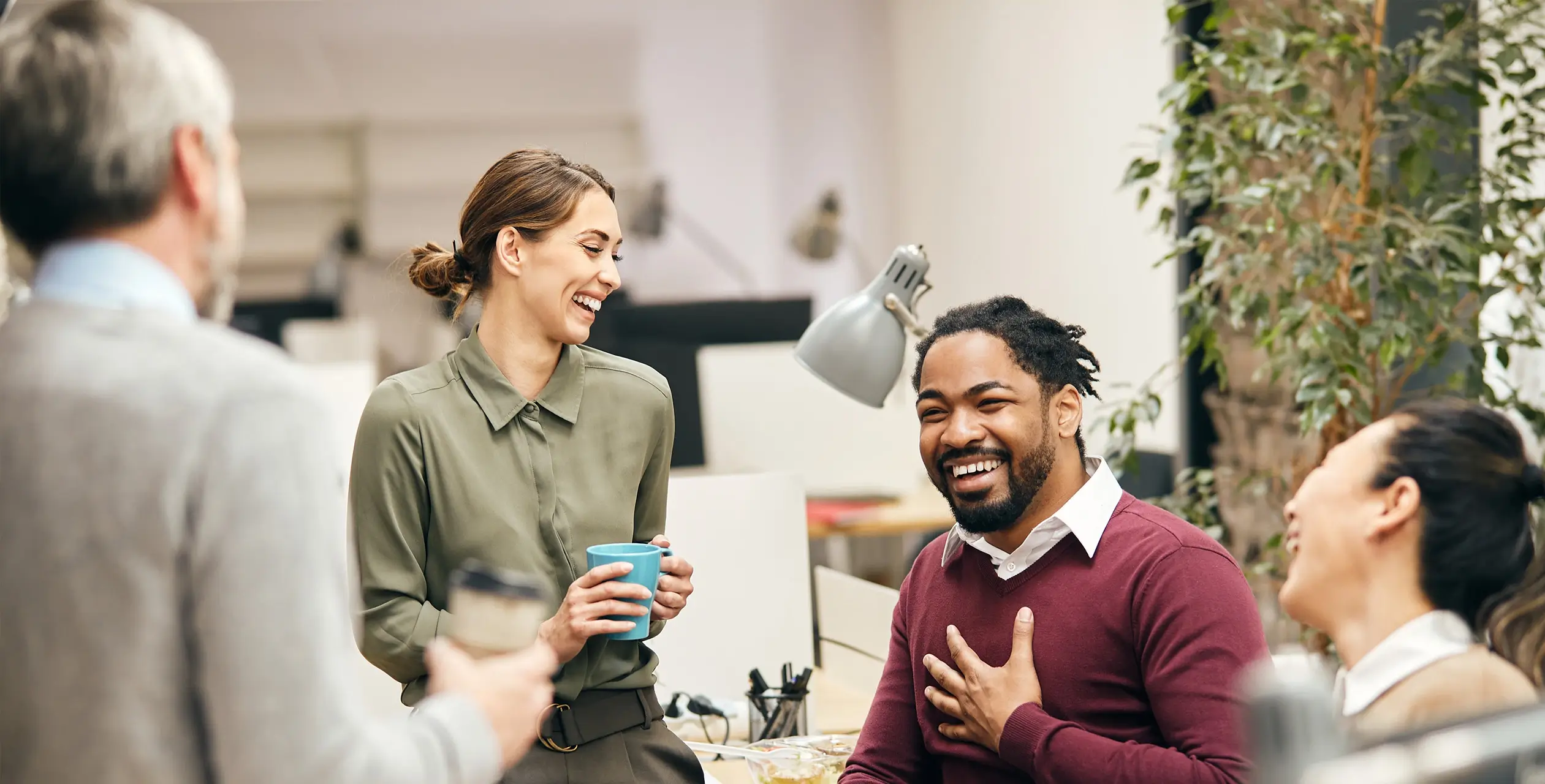 A group of people are gathered around a table, engaged in conversation. The setting is indoors, and they appear to be smiling and participating in an event related to their jobs. The image captures the diverse clothing styles of the individuals as they share ideas or stories.