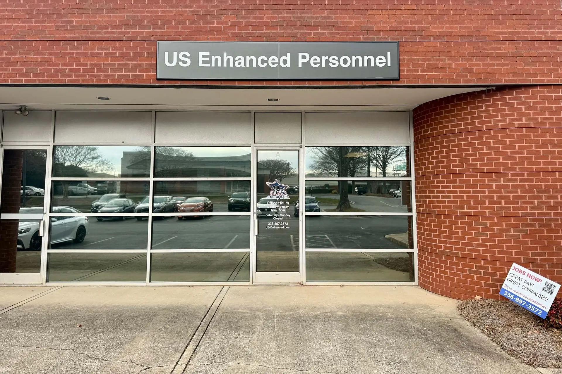 The image depicts a building featuring glass doors, likely serving as an entrance. There is a sign that reads "US Enhanced Personnel JONS NOW!" along with additional contact information. The setting appears to be outdoors, showcasing the storefront and its surroundings, including parked vehicles nearby.