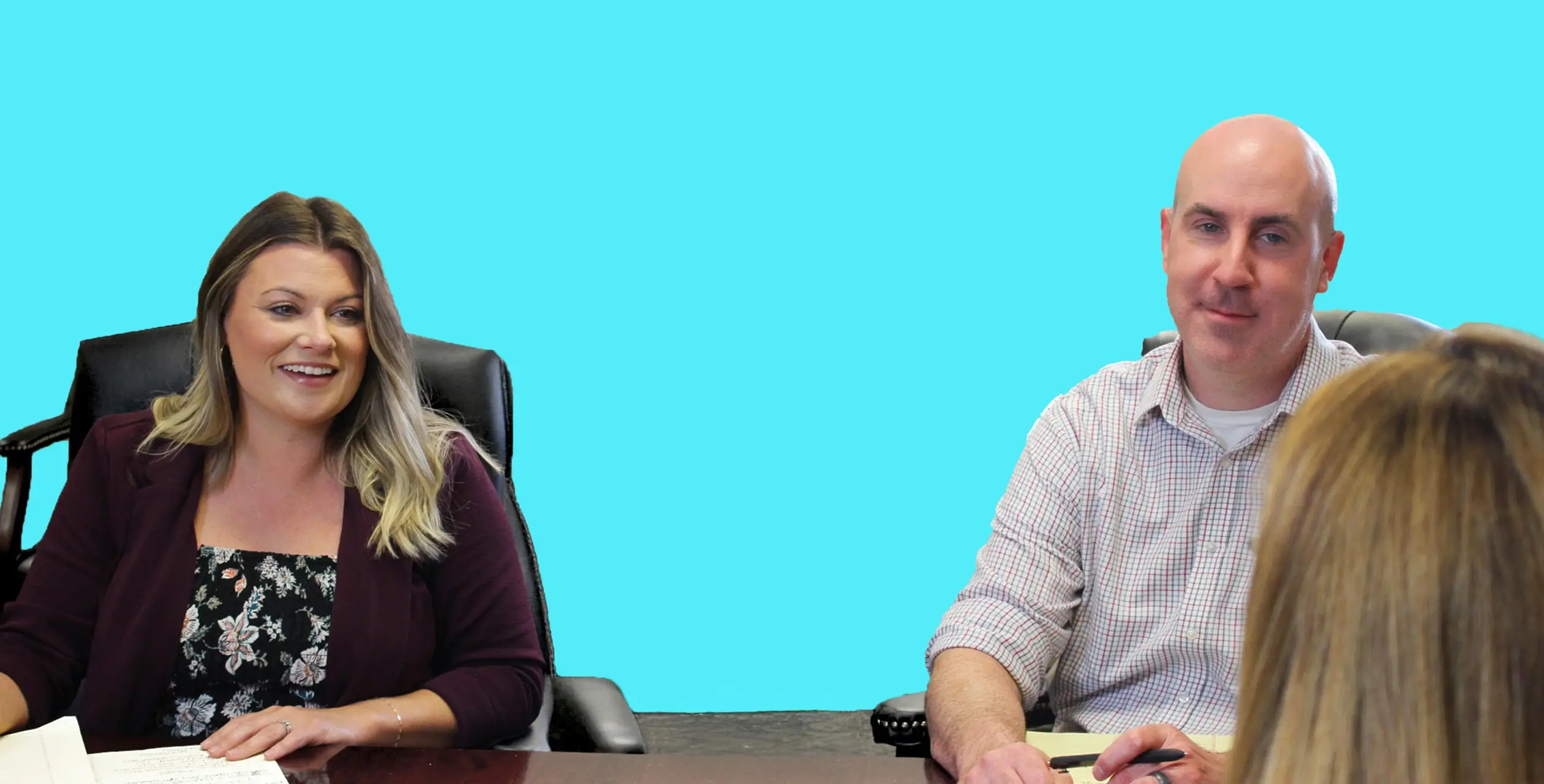 The photo shows three people, two of whom are sitting behind a desk. The woman on the desk's left side is smiling and wearing a floral top with a dark cardigan or jacket. The man on the desk's right is also smiling and dressed in a light-colored button-down shirt. The third person is out of focus in the foreground, with only the back of their head visible. The background appears to be a solid light blue color, possibly from a screen or wall.