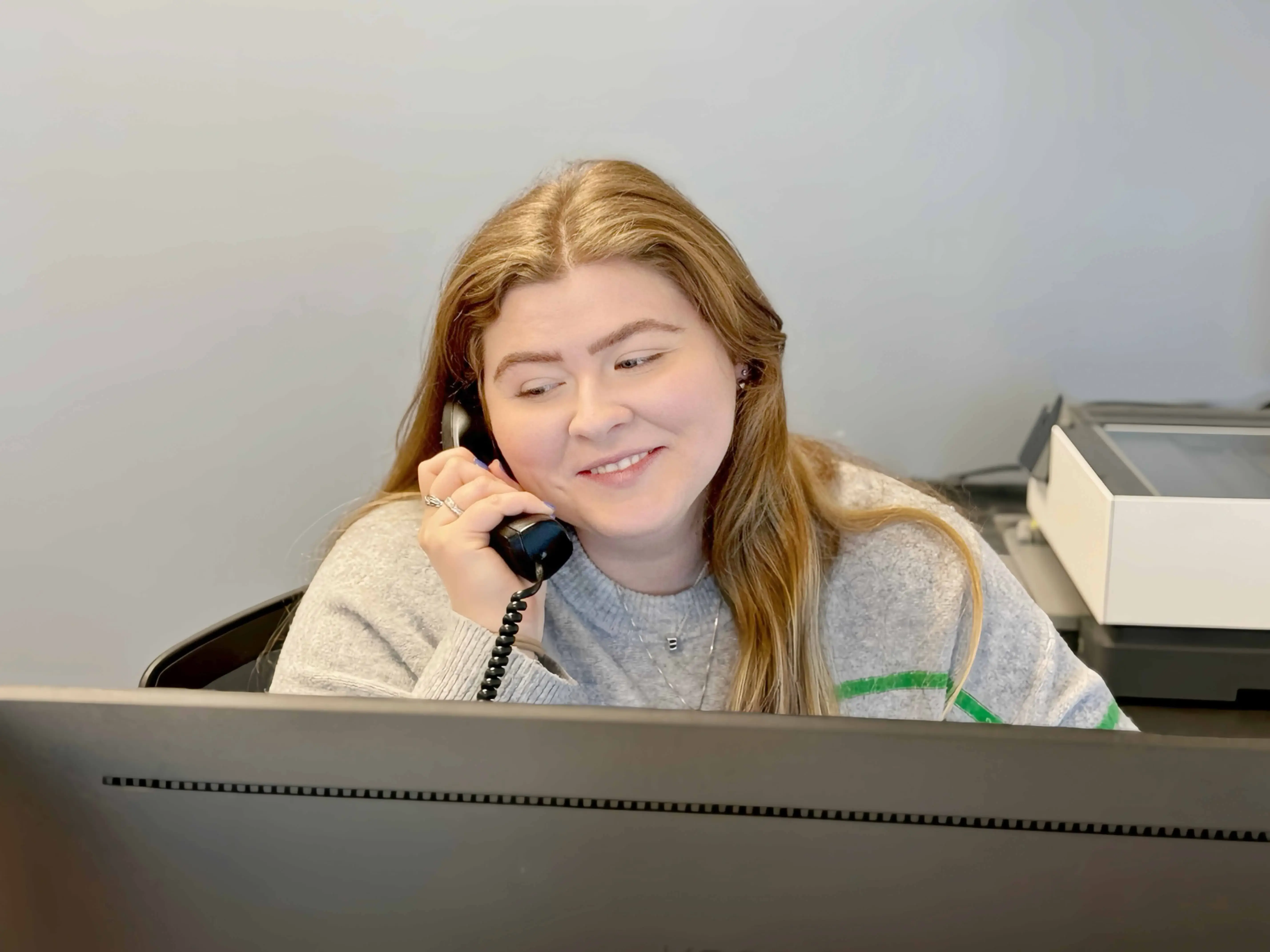 The image depicts a person sitting at a desk, engaged in a conversation on a traditional black telephone with a coiled cord, suggesting an older model or perhaps a preference for classic phones over digital options. The person is wearing a grey sweater adorned with what appears to be a green stripe down one of the arms or sleeves.

In front of this individual is a dark-colored monitor displaying some kind of graphical interface, possibly indicating the person is working on data analysis, graphic design, or programming work visible on the screen. The desk also houses other office essentials such as a white keyboard and what seems to be a closed notebook or planner with a pen attached, hinting at ongoing work or preparation for meetings.

Behind the seated figure is a partial view of a room with light walls and what could be a large piece of art or a decorative object, providing a minimalistic backdrop to the scene that suggests a professional setting. There's an element of natural light filtering through from the top left corner of the frame, illuminating the workspace slightly and contributing to the overall ambient feel of the photo.