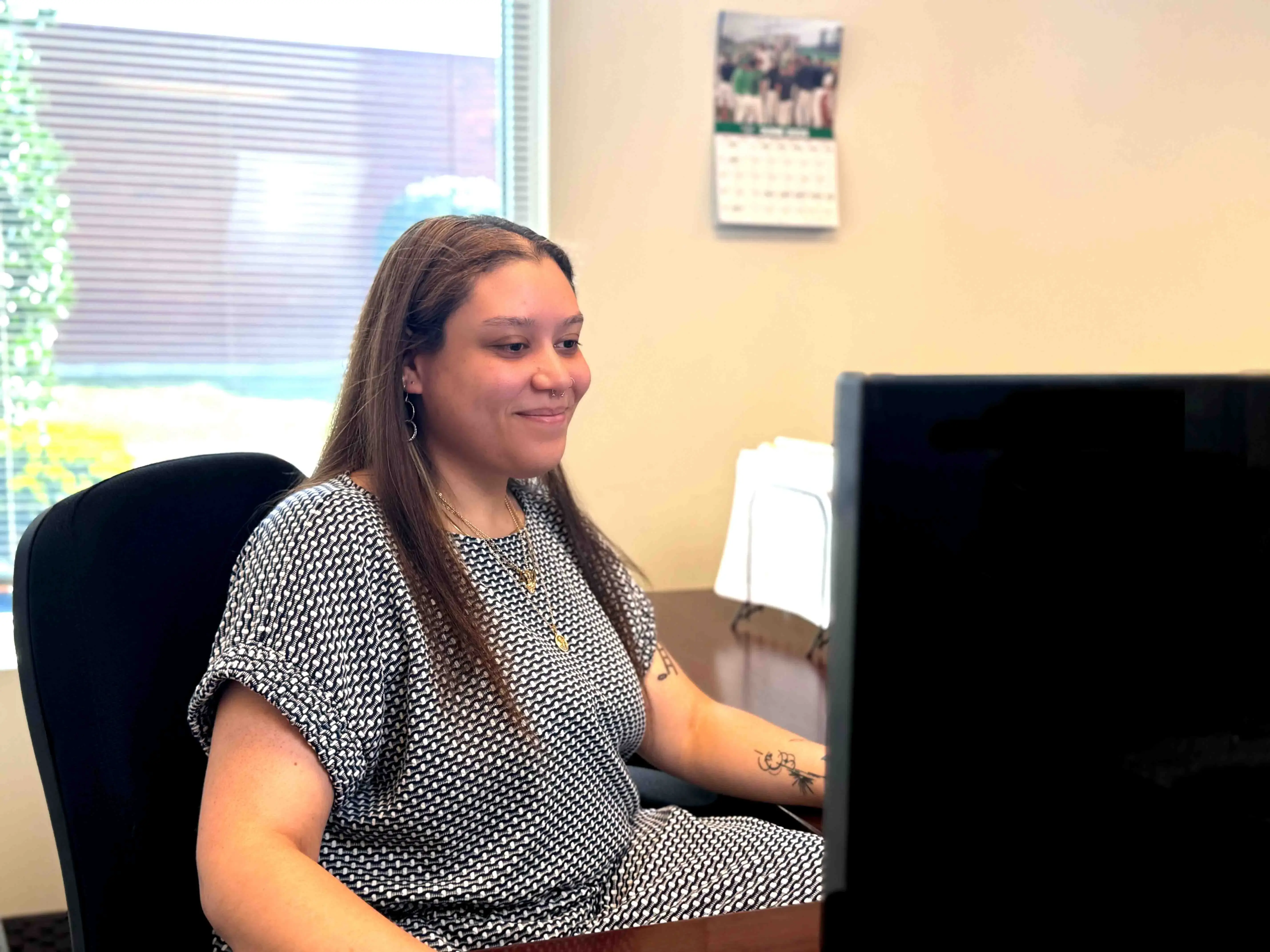 The image shows a person seated at a desk in an office environment, indicated by the presence of a large monitor displaying what appears to be data or a presentation, typical of a professional setting. The individual is wearing a short-sleeved shirt with a black and white geometric pattern.

In the background, there's a window that allows natural light to illuminate the space, with blinds partially closed. The daylight filters through the blinds, creating a dappled effect on the wall and highlighting the person's relaxed yet attentive posture. The person seems to be focused on the content on the screen before them, suggesting they are engaged in work.

Additionally, behind the chair and on the desk, there appear to be documents organized using a document holder, reinforcing the notion that this is a workspace used for tasks that require written materials or digital screens for reference or input.

There's also a mounted calendar on the wall, indicating that the environment is likely used for scheduled or time-managed activities, which could suggest a role that involves meetings, appointments, or deadlines.The general atmosphere of the scene conveys a sense of productivity and orderliness within the professional environment.