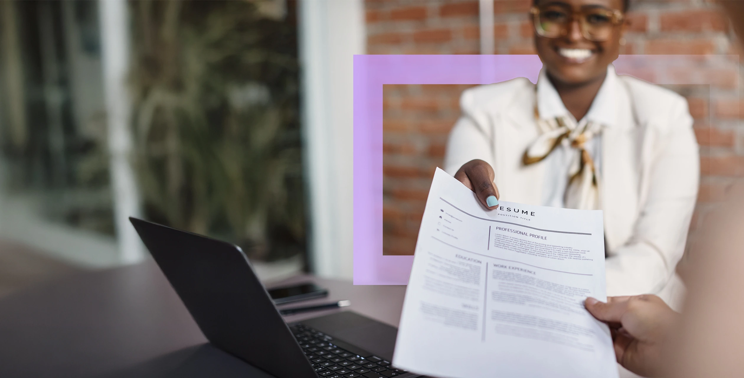 A woman is holding a piece of paper while seated at a laptop. She is indoors, and the setting includes a wall in the background. The image captures her engaged in an activity that involves using technology, reflecting elements of work or study.
