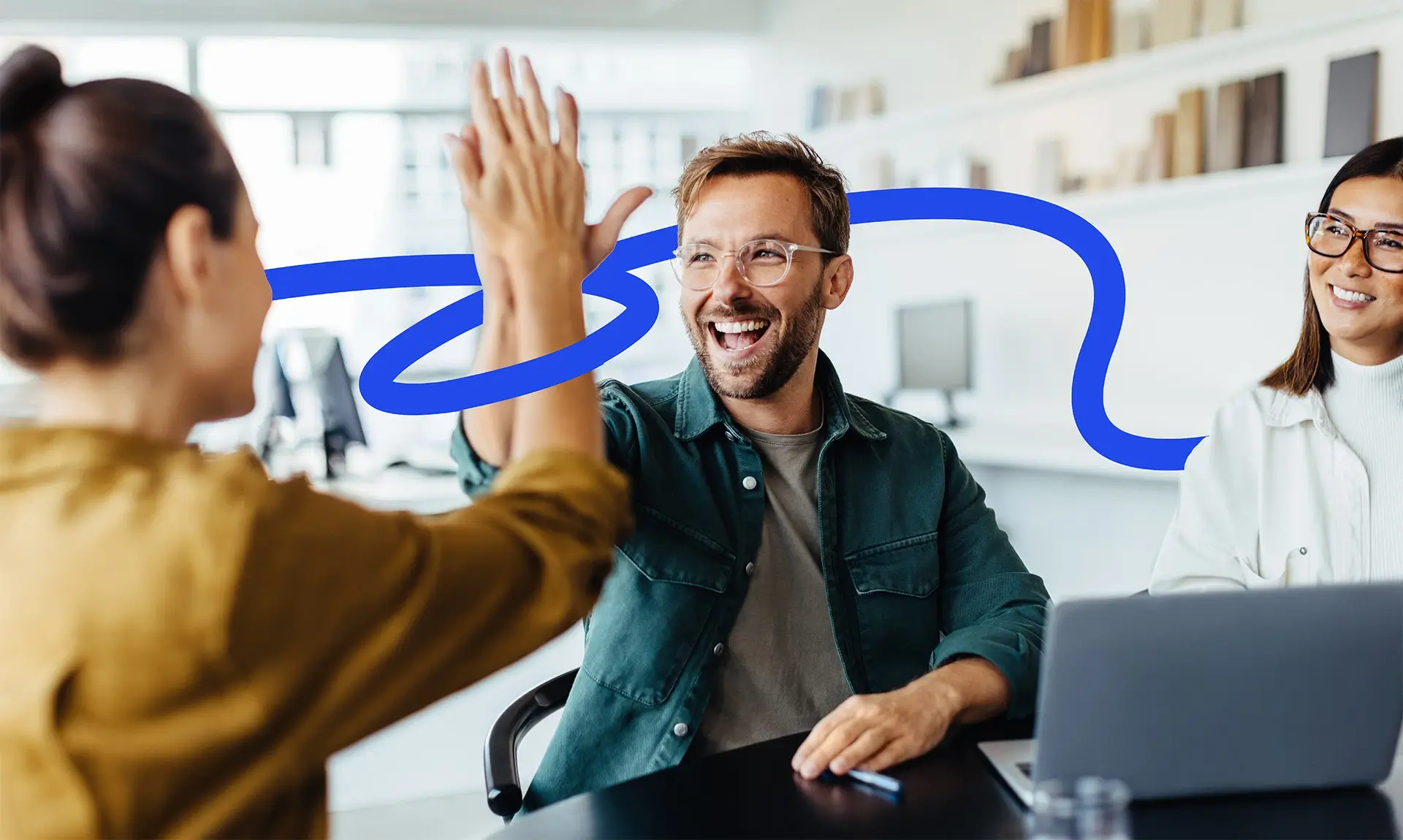 The image features two men indoors, both smiling as one gives a high five to the other. They are dressed in casual clothing and are engaged in a moment of celebration or camaraderie. A laptop and some office-related items may be visible in the background, suggesting a workplace environment.