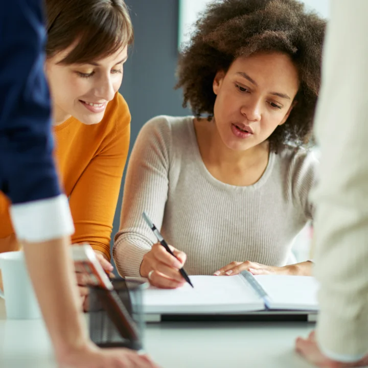 A group of individuals is gathered indoors, looking intently at a piece of paper. They appear to be engaged in a learning environment, suggesting an educational setting such as a classroom. The scene captures the focus and curiosity of students as they interact with the material presented.