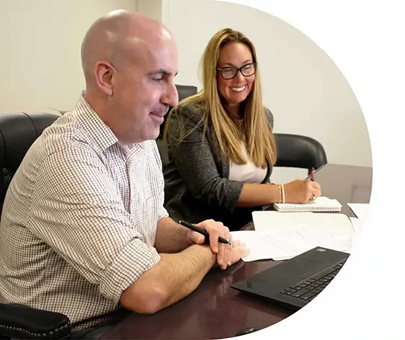 A man and woman are sitting at a table in an indoor office setting. They appear to be engaged in a discussion, smiling and collaborating over work-related matters. The environment includes typical office furniture and decor, indicating a professional atmosphere.
