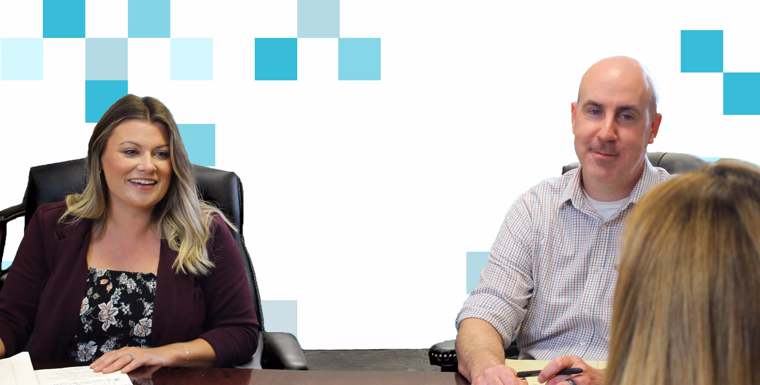 The photo shows three people, two of whom are sitting behind a desk. The woman on the desk's left side is smiling and wearing a floral top with a dark cardigan or jacket. The man on the desk's right is also smiling and dressed in a light-colored button-down shirt. The third person is out of focus in the foreground, with only the back of their head visible. The background appears to be a solid light blue color, possibly from a screen or wall.