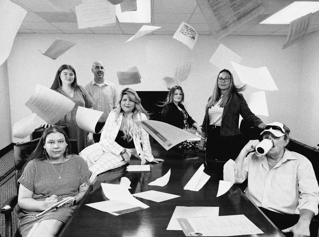 Group of people in a meeting room with papers flying in the air, some seated, others standing, creating a chaotic yet fun scene.