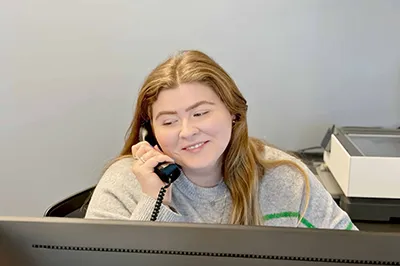 The image depicts a person sitting at a desk, engaged in a conversation on a traditional black telephone with a coiled cord, suggesting an older model or perhaps a preference for classic phones over digital options. The person is wearing a grey sweater adorned with what appears to be a green stripe down one of the arms or sleeves.

In front of this individual is a dark-colored monitor displaying some kind of graphical interface, possibly indicating the person is working on data analysis, graphic design, or programming work visible on the screen. The desk also houses other office essentials such as a white keyboard and what seems to be a closed notebook or planner with a pen attached, hinting at ongoing work or preparation for meetings.

Behind the seated figure is a partial view of a room with light walls and what could be a large piece of art or a decorative object, providing a minimalistic backdrop to the scene that suggests a professional setting. There's an element of natural light filtering through from the top left corner of the frame, illuminating the workspace slightly and contributing to the overall ambient feel of the photo.