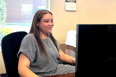 The image shows a person seated at a desk in an office environment, indicated by the presence of a large monitor displaying what appears to be data or a presentation, typical of a professional setting. The individual is wearing a short-sleeved shirt with a black and white geometric pattern.

In the background, there's a window that allows natural light to illuminate the space, with blinds partially closed. The daylight filters through the blinds, creating a dappled effect on the wall and highlighting the person's relaxed yet attentive posture. The person seems to be focused on the content on the screen before them, suggesting they are engaged in work.

Additionally, behind the chair and on the desk, there appear to be documents organized using a document holder, reinforcing the notion that this is a workspace used for tasks that require written materials or digital screens for reference or input.

There's also a mounted calendar on the wall, indicating that the environment is likely used for scheduled or time-managed activities, which could suggest a role that involves meetings, appointments, or deadlines.The general atmosphere of the scene conveys a sense of productivity and orderliness within the professional environment.