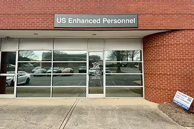 The image depicts a building featuring glass doors, likely serving as an entrance. There is a sign that reads "US Enhanced Personnel JONS NOW!" along with additional contact information. The setting appears to be outdoors, showcasing the storefront and its surroundings, including parked vehicles nearby.