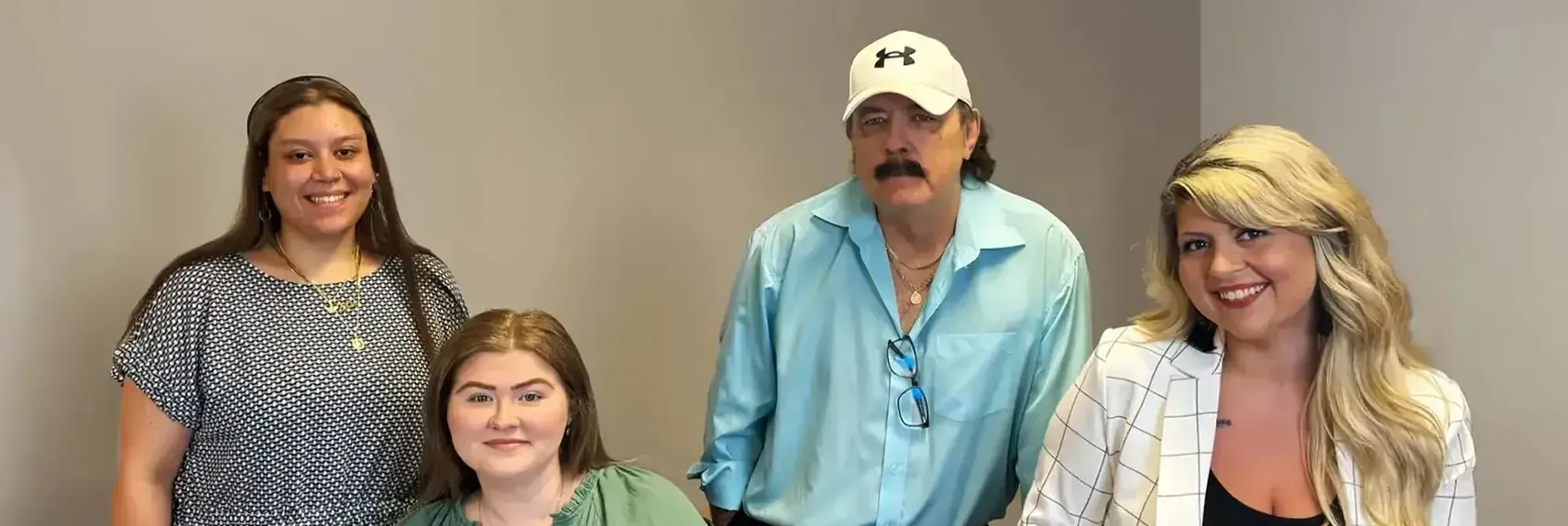 Four people posing indoors against a plain wall, smiling at the camera—two women standing on the left and right, one woman seated in front, and a man wearing a cap and button-down shirt standing in the back.