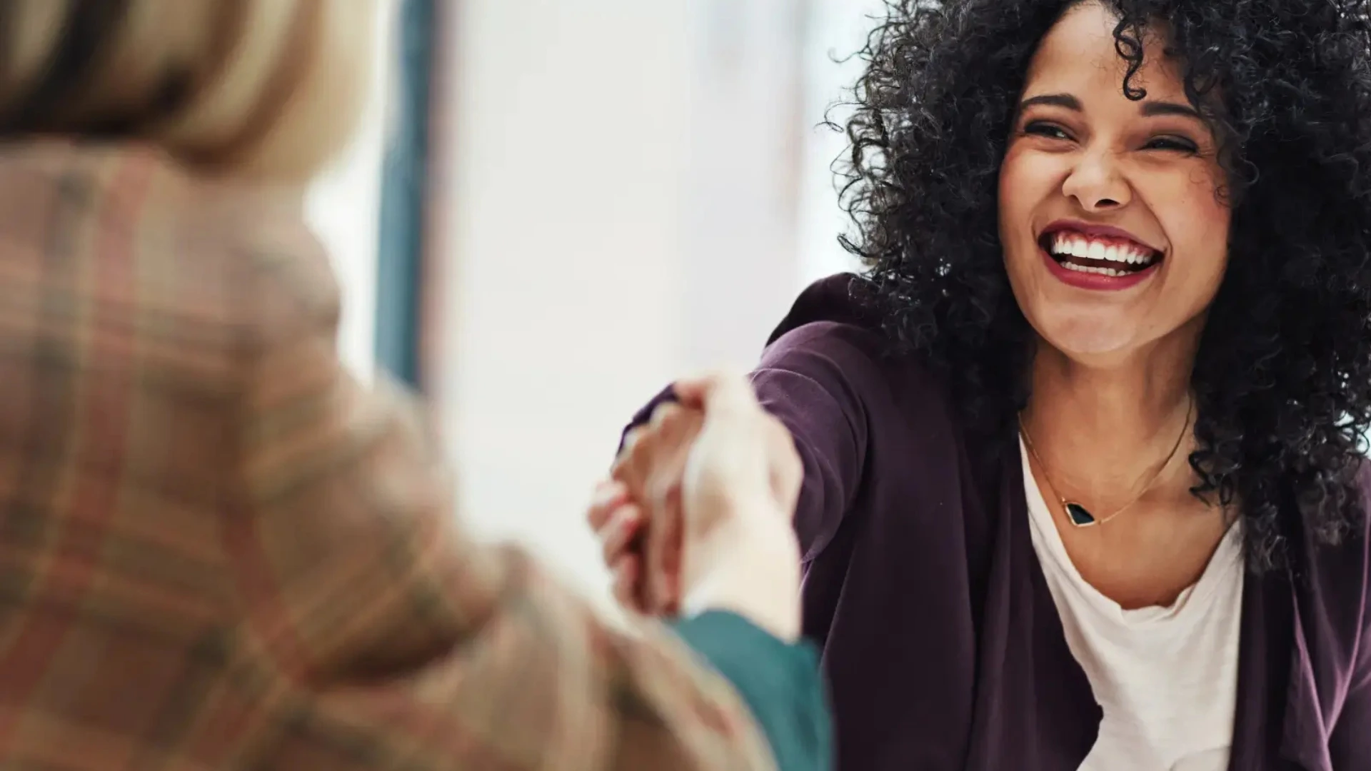 This image shows a close-up of two people greeting each other with a handshake. In the foreground, a woman with blonde hair, dressed in a plaid blazer, is extending her hand. In the background, a woman with curly dark hair, wearing a white top and dark blazer, is smiling brightly while shaking hands. The focus is on the smiling woman, capturing her warm expression and eye contact, while the background remains softly blurred. The overall tone suggests professionalism, friendliness, and positive connection, making it well-suited for business or workforce-related themes.