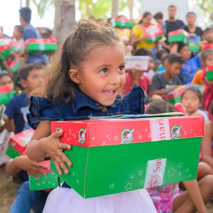 The image shows a young girl smiling brightly while holding a green and red shoebox gift. She looks excited and joyful. The box is part of a larger distribution, as many other children in the background are also holding similar gift boxes. The scene suggests a charitable event or celebration where children are receiving presents, and the girl’s expression captures the happiness of the moment.
