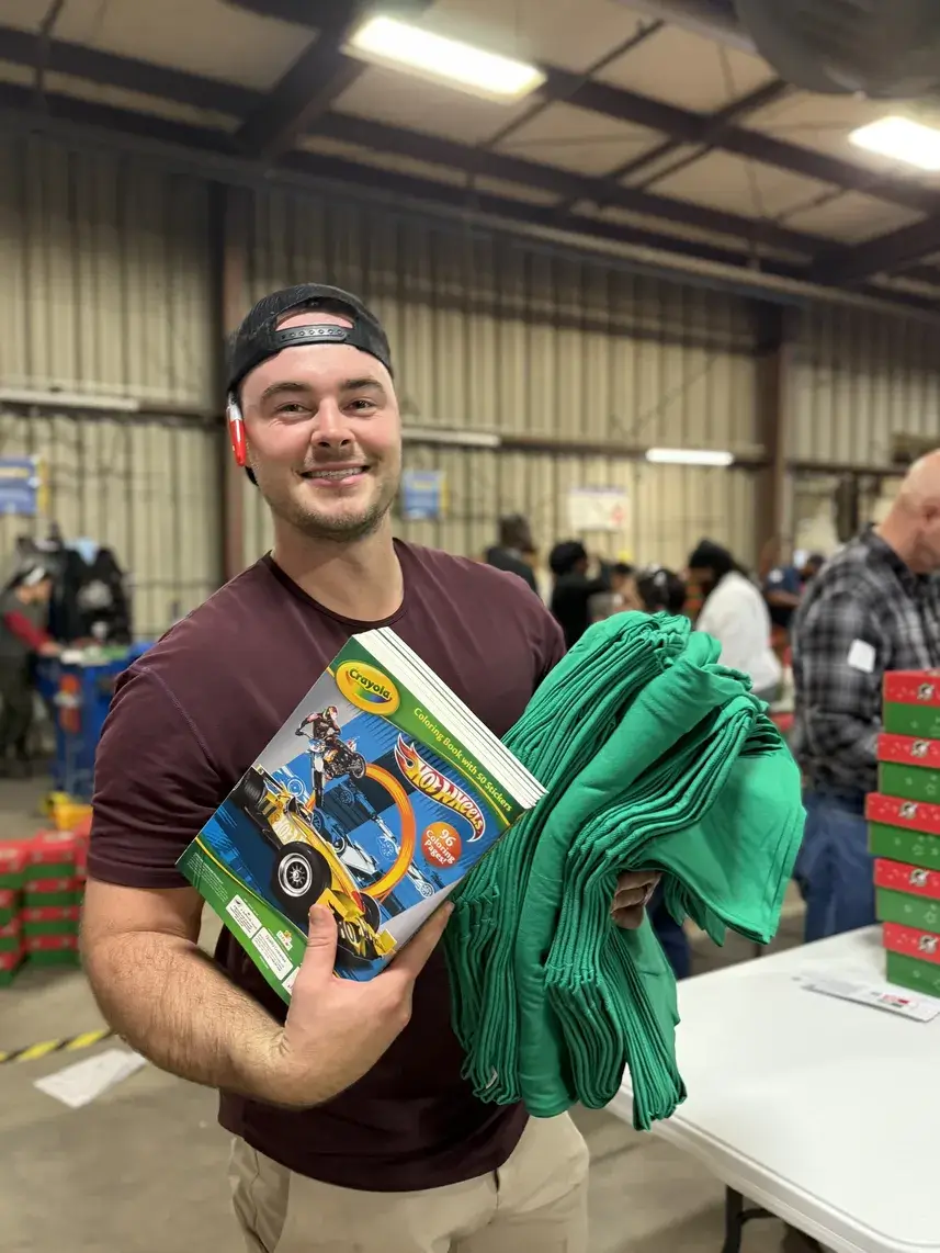 The image shows a smiling man wearing a backward baseball cap, standing indoors in what looks like a warehouse or packing facility. He is holding a Crayola Hot Wheels coloring and activity pad in one arm and a stack of folded green cloths or T-shirts in the other. In the background, people are busy working, and there are stacks of green-and-red shoeboxes, commonly associated with Operation Christmas Child, visible on the table and in the distance. The scene suggests he is participating in a volunteer event to prepare and pack shoebox gifts for children.