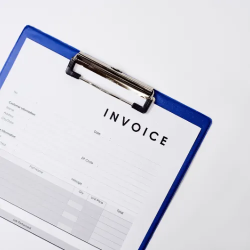 A close-up of a clipboard holding a paper invoice. The word INVOICE is printed clearly at the top in bold letters, and the rest of the document has structured fields for details such as contact information, date, and payment items. The clipboard is blue with a metal clip.
