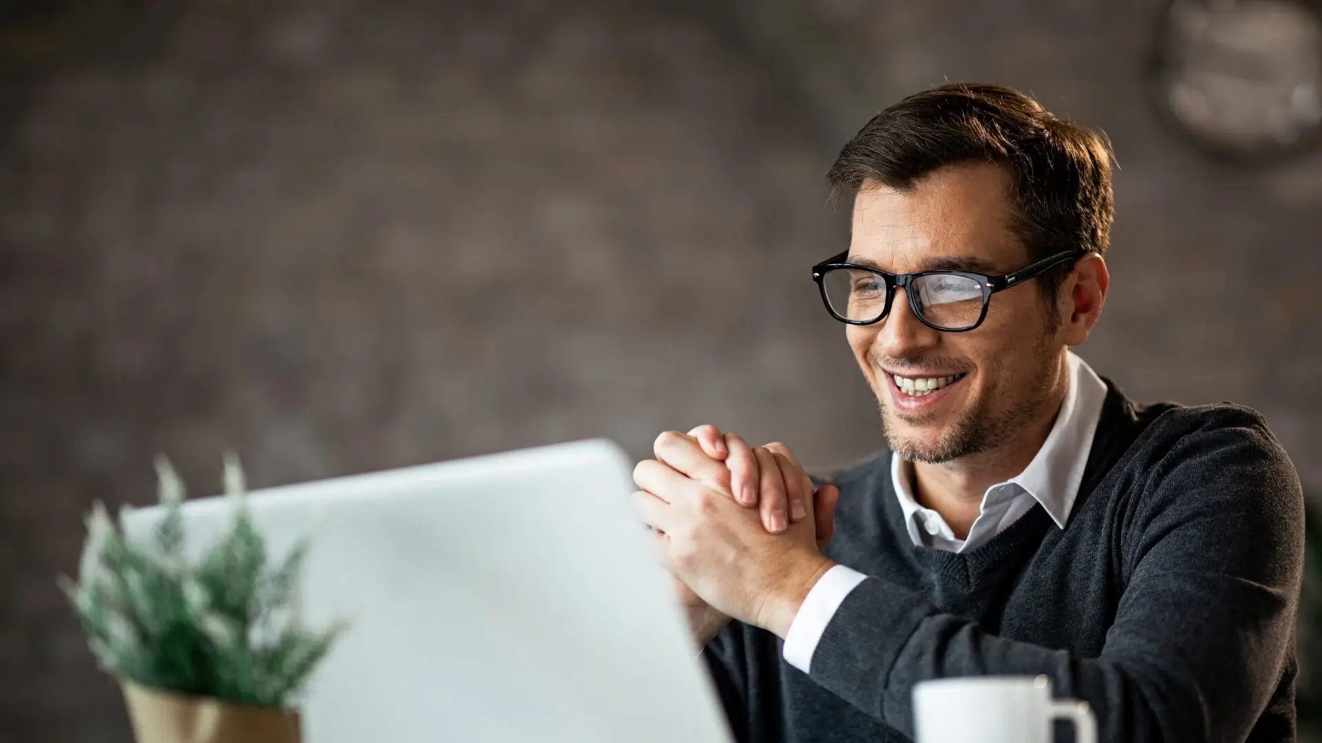 He appears to be indoors, and he is wearing glasses. The background features a wall, and his positive expression suggests he is engaged with something enjoyable on the computer.