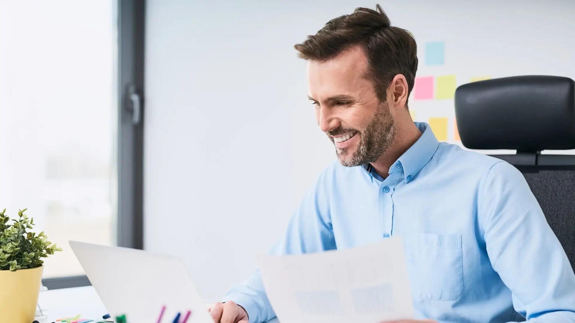 Smiling man in a blue shirt working at a desk with a laptop and paper in an office.