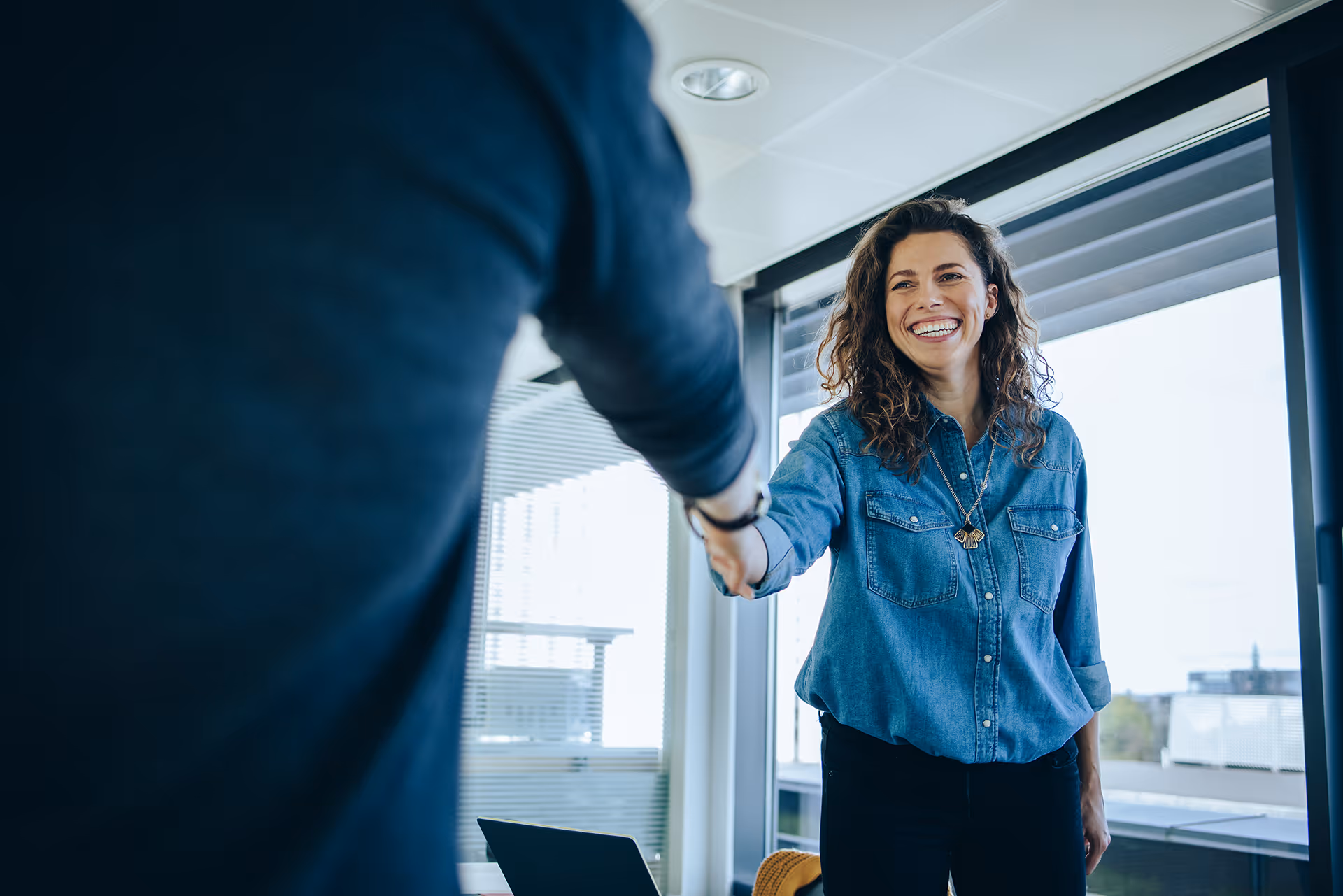 Smiling woman in glasses and white blazer shaking hands with a man in an office setting.