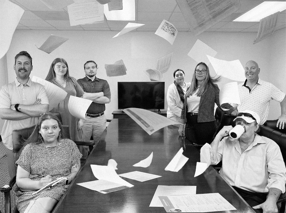 Seven people posing in a conference room with papers flying in the air around a long table.