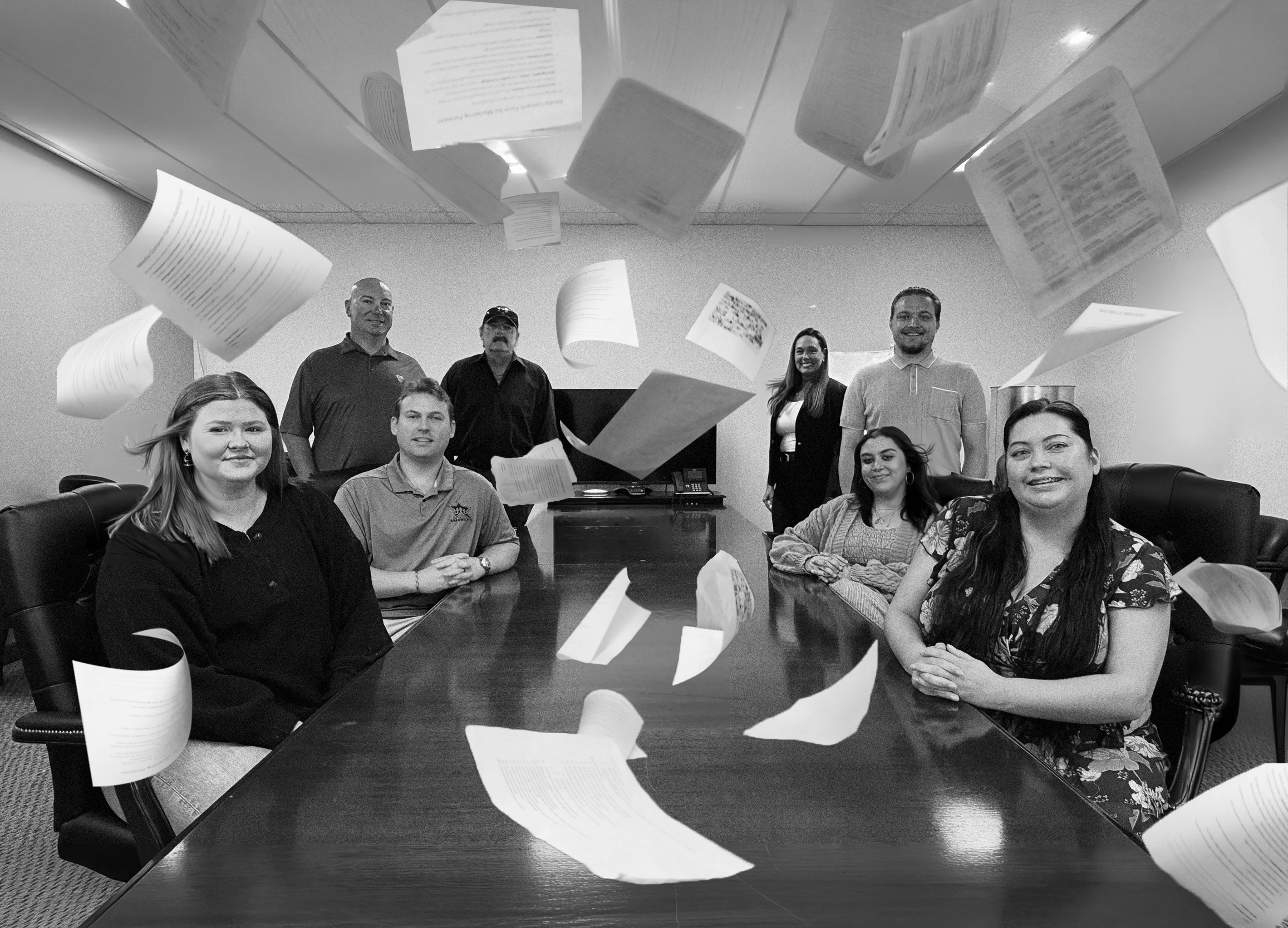 Group of people sitting and standing around a conference table with papers flying in the air.