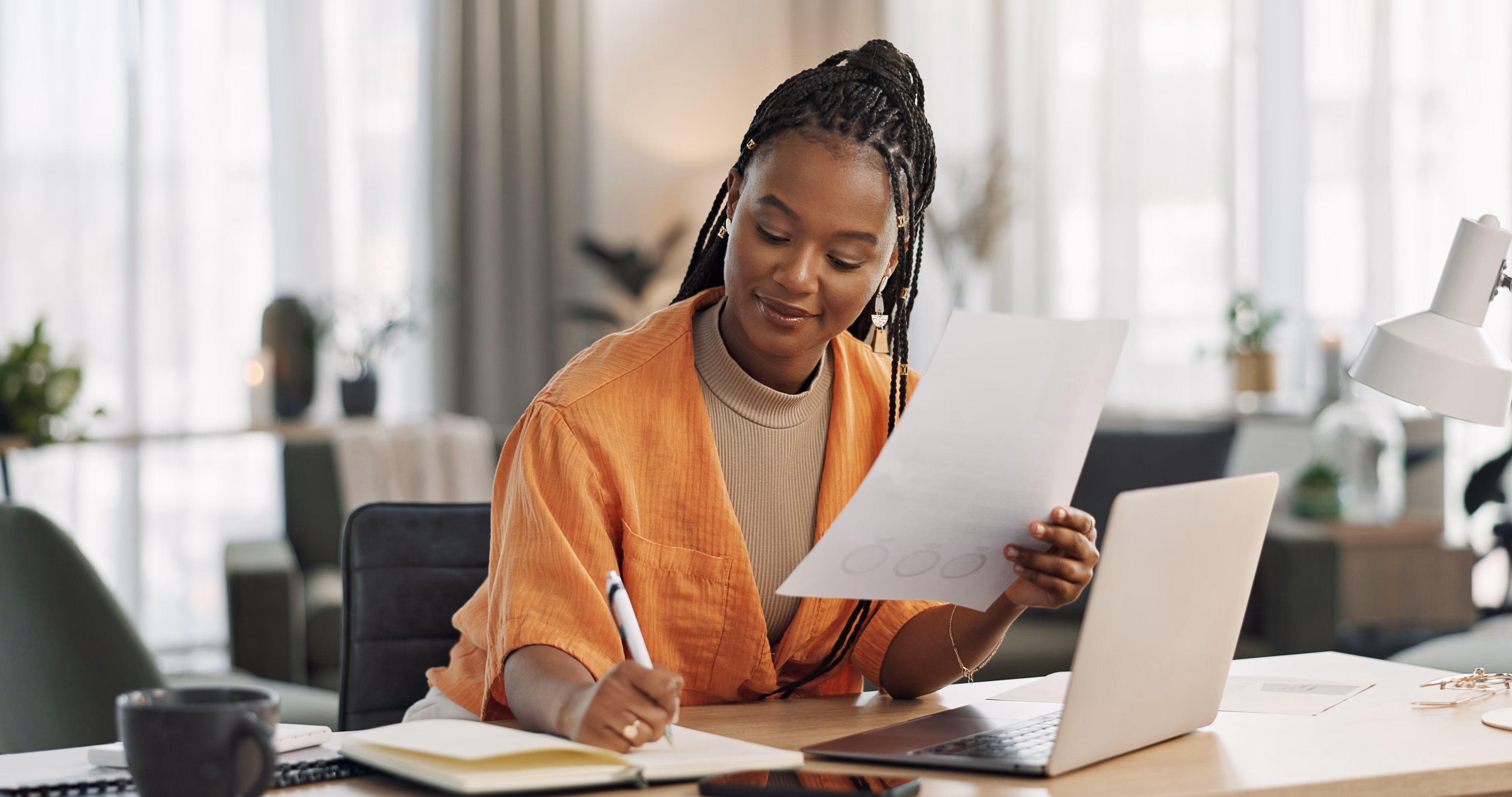 Woman in an orange shirt writing in a notebook while holding a paper and sitting at a desk with a laptop.