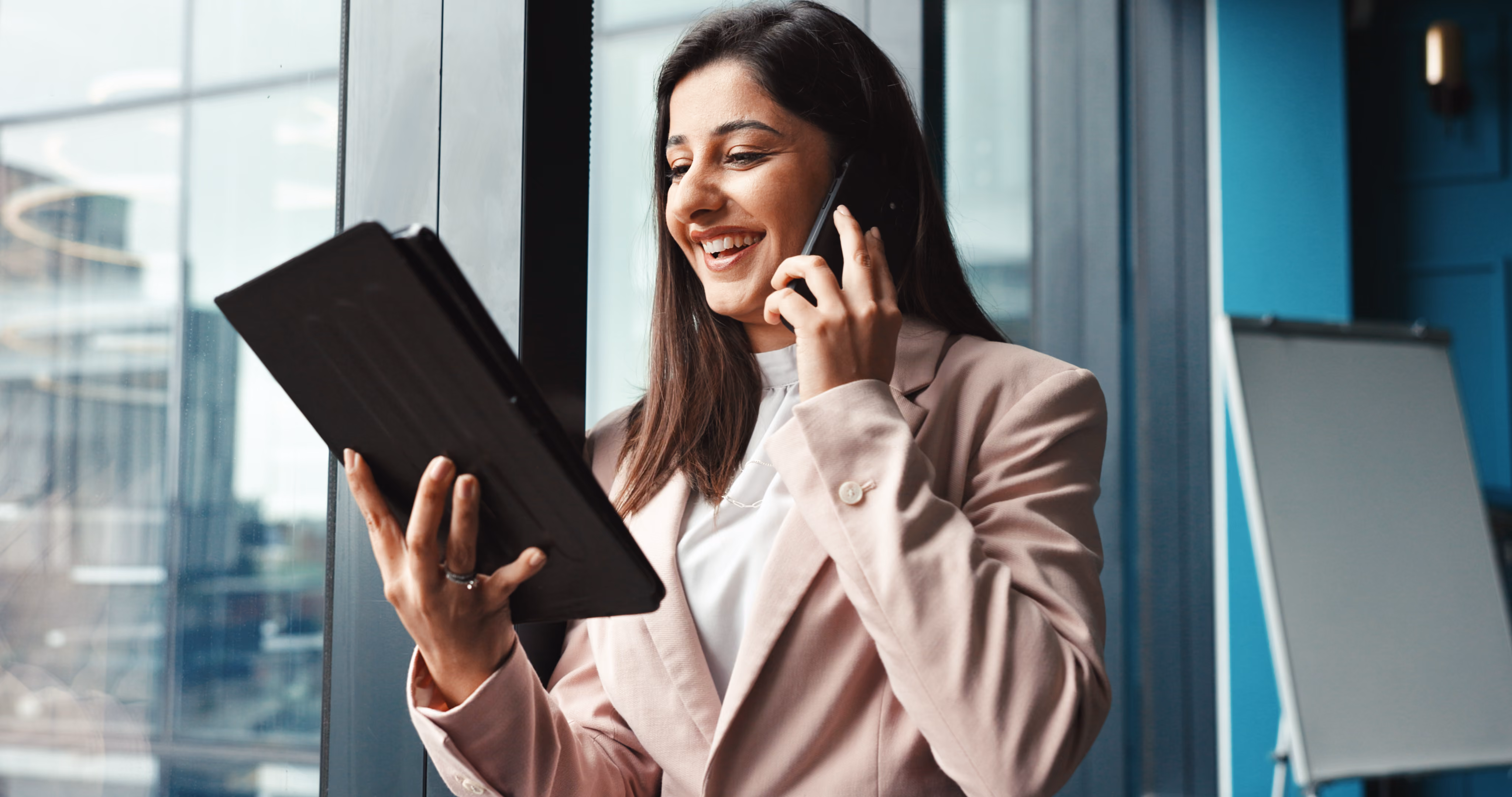 Smiling woman in a blazer talking on phone and holding a tablet near an office window.