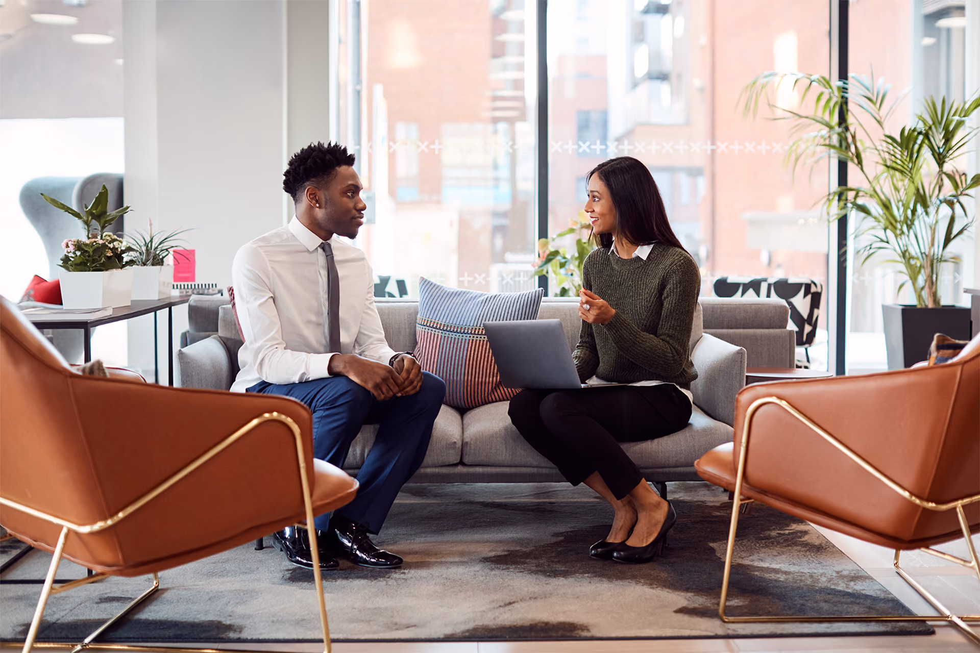 Two people sitting on a sofa in a modern office having a discussion, one holding a laptop.