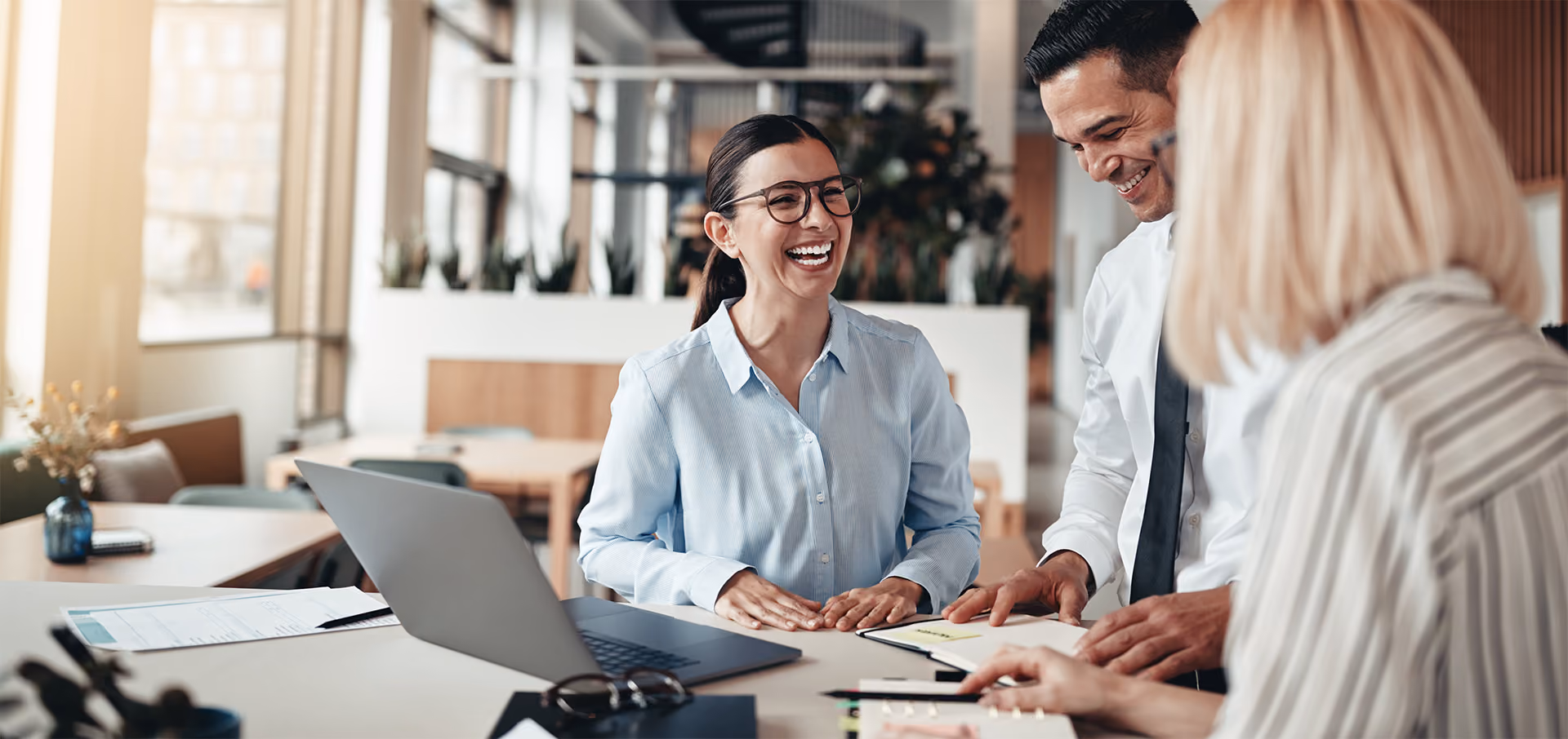 Three colleagues smiling and working together at a desk with a laptop and documents in a bright office.