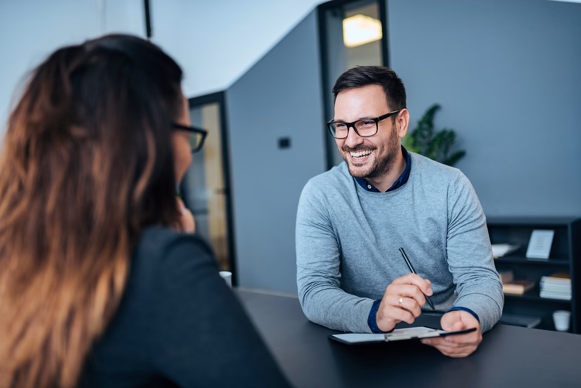 Smiling man with glasses holding a pen and clipboard during a discussion with a woman in an office.