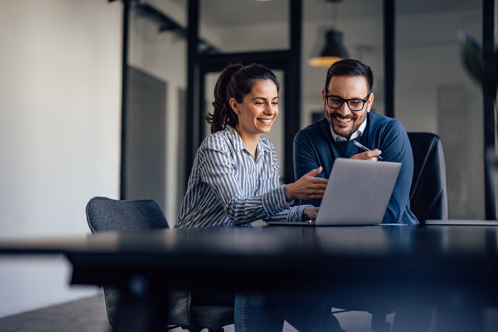 Smiling man and woman looking at a laptop, discussing something in an office setting.