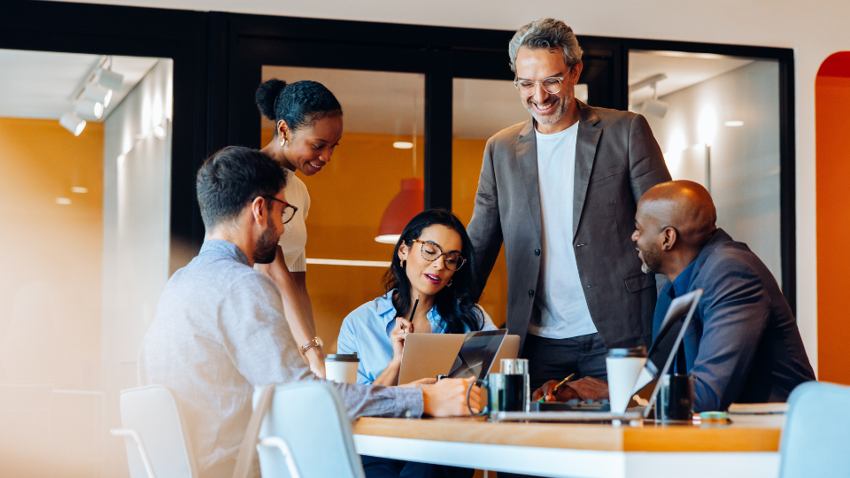 Five diverse professionals collaborating and smiling around a table with laptops and coffee cups in an office.